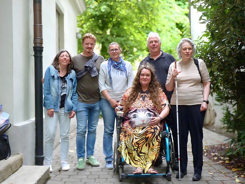 Eine Gruppe von sieben Personen steht in einer schmalen Gasse im Freien, umgeben von hellen Gebäuden und grüner Vegetation. In der Mitte sitzt eine Frau im Rollstuhl, sie hat eine große Tasche auf dem Schoß, die wie ein Katzenkopf aussieht. Die anderen sechs Personen stehen um sie herum, zwei Männer und vier Frauen. Eine der Frauen rechts außen hält einen weißen Blindenstock. Alle blicken in die Kamera und lächeln. Die Atmosphäre wirkt freundlich und entspannt.